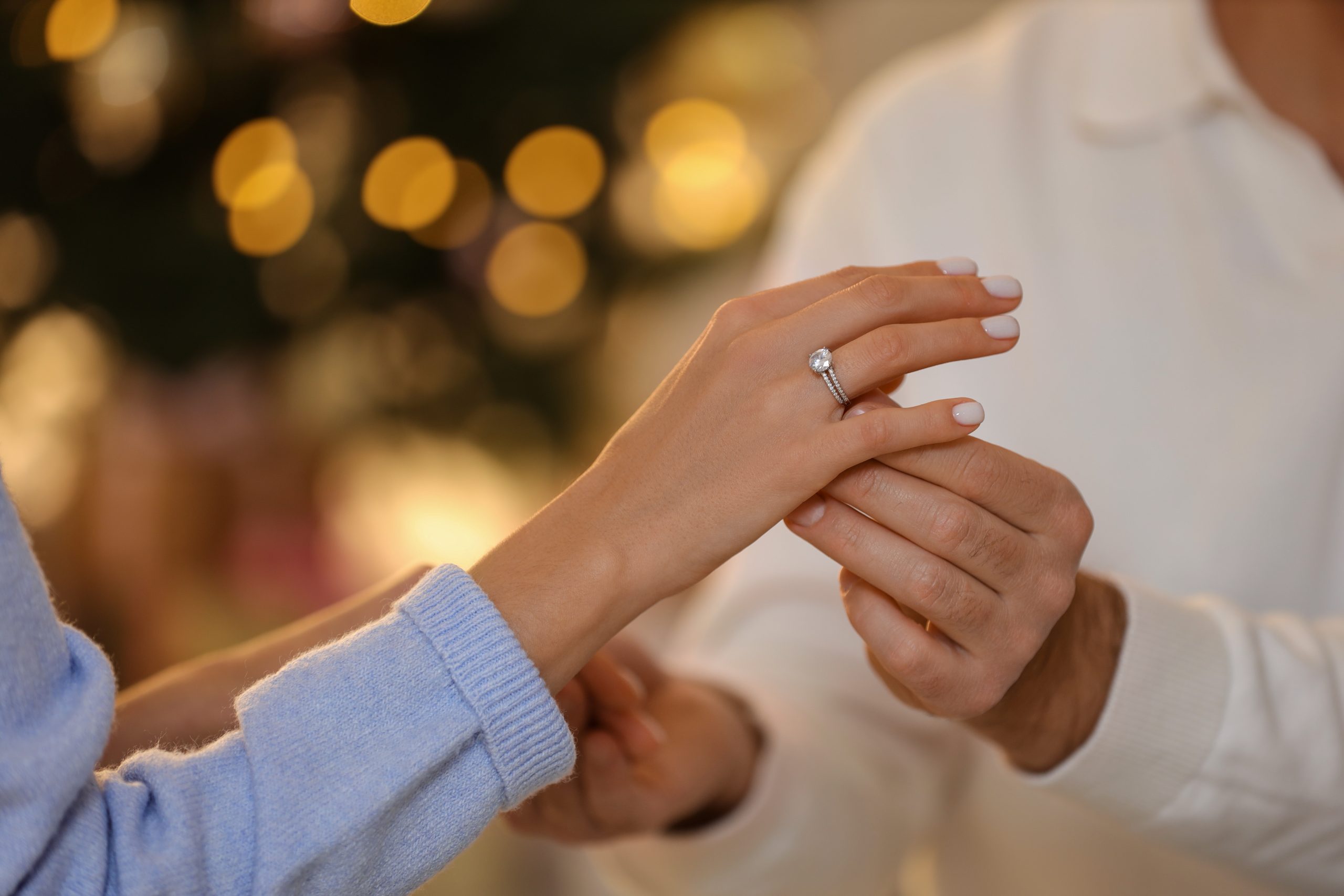Upper body of man and woman touching hands in front of lights in the background the woman wears a ring on her ring finger