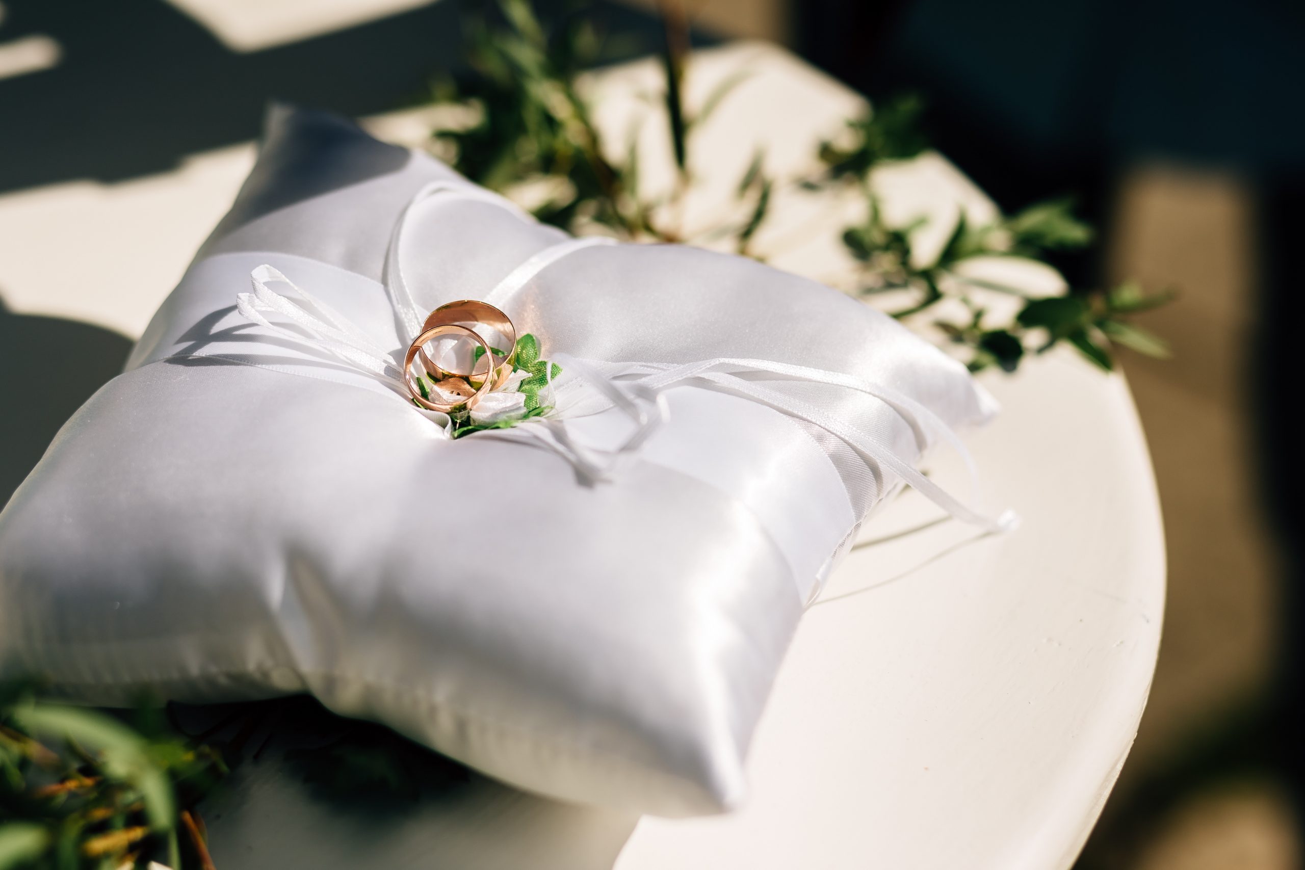 two golden rings on a white ring cushion with flowers on a white table