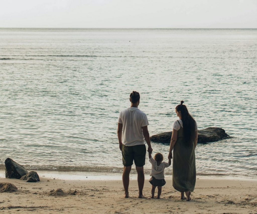 Paar mit Kleinkind am Strand, Blick aufs Meer, symbolisiert Familie und Freizeit.